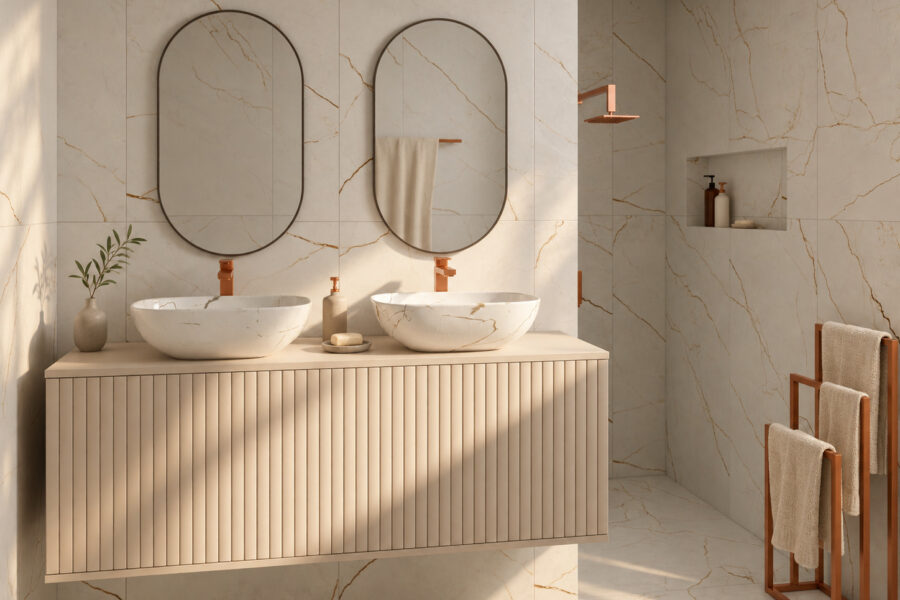 Bathroom in warm daylight: a wall-hung vanity with one fluted cashmere front, two marble-veined countertop basins with copper taps under two oval mirrors, a walk-in shower with a copper shower head and a copper towel stand.